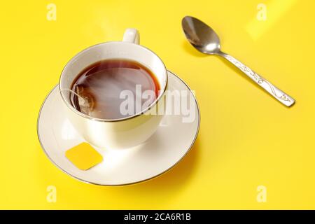 Morgensonne scheint bei frisch gebrühter dampfender Tasse Tee auf gelbem Brett, Beutel noch hinein, silberner Löffel daneben Stockfoto