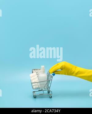 Weibliche Hand in einem gelben medizinischen Handschuh hält einen Spielzeug-Einkaufswagen mit Medikamenten auf blauem Hintergrund. Hygiene im Laden. Coronavirus-Pandemie, covid Stockfoto