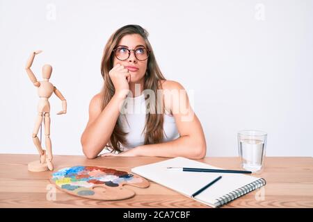 Schöne junge kaukasische Frau Künstler Maler sitzen auf Schreibtisch Denken konzentriert über Zweifel mit Finger auf das Kinn und schauen bis wundernd Stockfoto