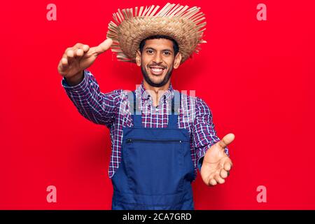 Junger lateinischer Mann trägt Bauernhut und Schürze Blick auf die Kamera lächelnd mit offenen Armen für Umarmung. Fröhlicher Ausdruck umarmen Glück. Stockfoto