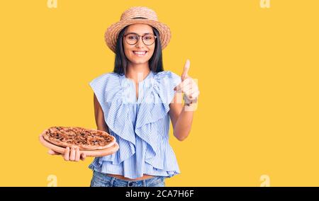 Junge schöne lateinische Mädchen trägt Sommerhut und Gläser Pizza halten glücklich und positiv lächelnd, Daumen nach oben tun ausgezeichnet und Genehmigung Zeichen Stockfoto