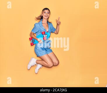 Junge schöne Mädchen halten Skate lächelnd glücklich. Springen mit Lächeln auf Gesicht tun Sieg Zeichen über isolierten gelben Hintergrund Stockfoto