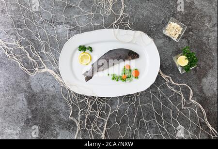 Eine ganze Regenbogenforelle auf einer weißen Servierplatte mit weißem Fischernetz mit Zitronen, Petersilie, Erbsen und Karotten auf einem grauen Marmortisch Stockfoto