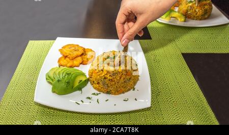 Nahaufnahme der Hand, die ein Krabbenbein von einem Teller mit Reis mit Krabben, begleitet von Avocado und Pataconen, auf einem weißen Teller auf einem Holztisch nimmt Stockfoto