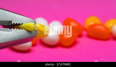 Eine Nahaufnahme eines Edelstahl-Zange Kopf Zerkleinern eine gelbe Jelly Bohne Lutscher oder Süßigkeiten. Leuchtend rosa lila verschwommen Hintergrund mit mehreren orange an Stockfoto