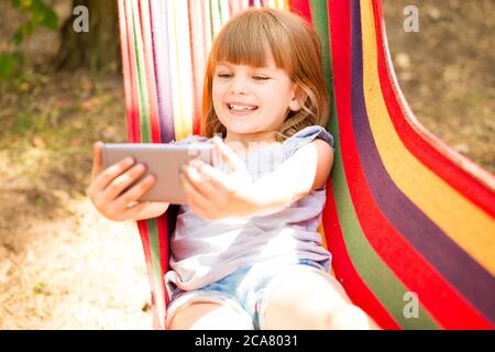 Nettes kleines Mädchen zu Fuß im Freien und Ruhe auf Hängematte im Sommerwald unter Selfie oder machen Videoanruf mit Handy. Stockfoto