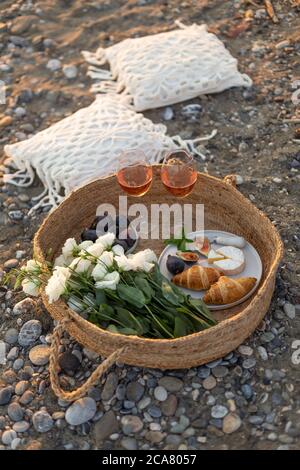 Vertikales Bild eines Strandpicknicks mit Croissants, Feigen, Kambrauner Käse, Rosenwein und weißen Lisianthus Blumen in einem gewebten Korb. Stockfoto