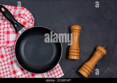 Leere schwarze Bratpfanne oder Pfanne mit Pfeffer und Meersalz auf einer rot karierten Serviette auf schwarzem Tisch. Draufsicht. Speicherplatz kopieren Stockfoto