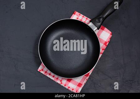 Leere schwarze Bratpfanne oder Pfanne auf einer rot karierten Serviette auf schwarzem Tisch. Draufsicht. Speicherplatz kopieren Stockfoto