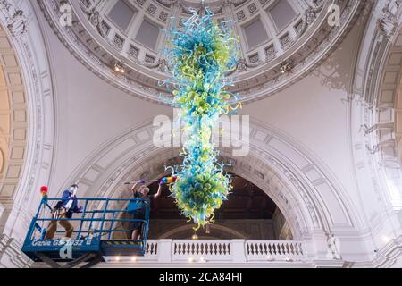 Museumstechniker Andy Monk reinigt den 27 Meter langen Rotunda-Kronleuchter von Dale Chihuly, der aus 1,300 blauen und grünen Glaselementen besteht, im Victoria and Albert Museum in London, während er sich auf die Wiedereröffnung am 6. August vorbereitet. Stockfoto