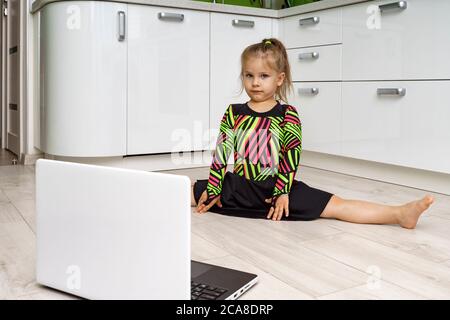 Kleines Mädchen 4 Jahre alt in einem Gymnastiktrikot ist in Online-Gymnastik zu Hause beschäftigt und schaut auf den Laptop. Sitzen auf einer Schnur Stockfoto