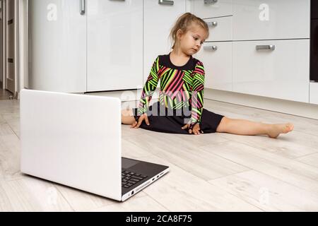 Kleines Mädchen in einem Turnanzug ist in Online-Gymnastik zu Hause beschäftigt und schaut auf einen Laptop. Sitzen auf einer Schnur Stockfoto