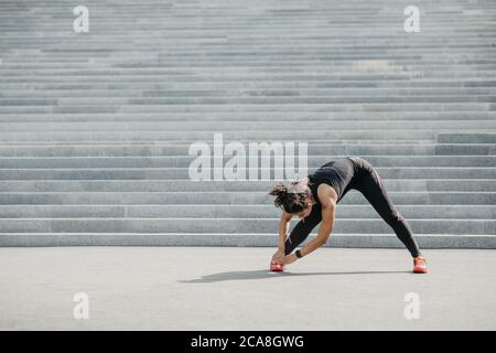 Dehnung nach dem Training. Afroamerikanische Frau mit Fitness-Tracker und kabellosen Kopfhörern macht Übungen für Beine Stockfoto