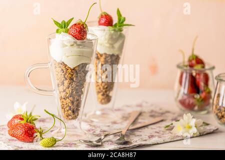 Zwei Gläser gesundes Frühstück mit Müsli und griechischem Joghurt, dekoriert mit Minze und roher Erdbeere, serviert auf weißem Holzhintergrund. Stockfoto