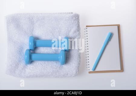 Gesundheits-Regime und Fitness-Symbole. Hanteln aus blauem Kunststoff liegen auf weißem Hintergrund auf einem Handtuch. Passform Form und Sportkonzept. Langhanteln neben leerem Copybook und cyanfarbenem Stift, Draufsicht Stockfoto