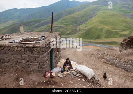 Xinaliq / Aserbaidschan - 8. Juli 2019: Frau mit Kopftuch arbeitet Wolle von Hand mit schönen Kaukasus-Bergen im Hintergrund zu rutschen Stockfoto