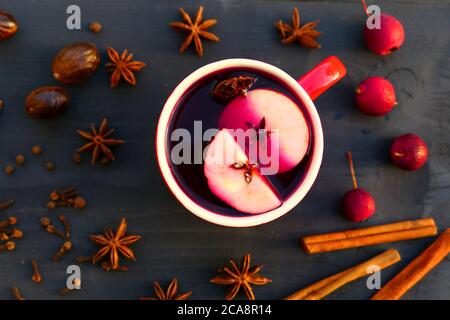 Weihnachtlicher Glühwein mit Gewürzen und Apfel in rotem Becher auf Holzhintergrund. Zimt, Muskatnuss, Sternanis, Nelken. Stockfoto