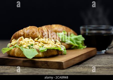 Croissant gefüllte Rühreier auf Holztablett mit heißem Kaffee Hintergrund. Stockfoto