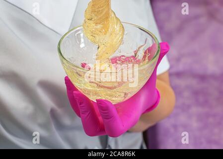 Meister der Enthaarung in rosa Latexhandschuhen zieht eine Holzstäbchen-Zuckerpaste aus der Schüssel Stockfoto