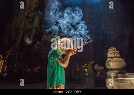 Kleiner Junge, der in einem buddhistischen Tempel betet, der Weihrauch hält Huyen Khong Höhle mit Schreinen, Marmorberge, Vietnam Stockfoto