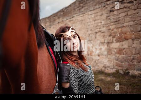 Ein schönes Mädchen steht neben einem Pferd vor dem Hintergrund einer mittelalterlichen Festung. Fantasy Kostüm, Kettenpost, Mantel, Krone. Eine Frau in einem magischen c Stockfoto