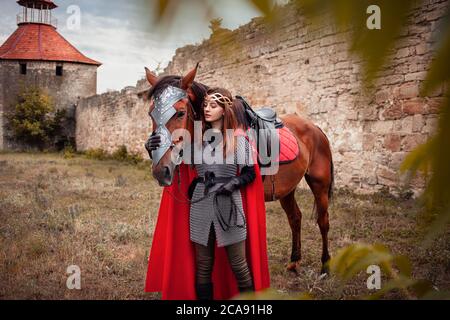 Ein schönes Mädchen steht neben einem Pferd vor dem Hintergrund einer mittelalterlichen Festung. Fantasy Kostüm, Kettenpost, Mantel, Krone. Eine Frau in einem magischen c Stockfoto