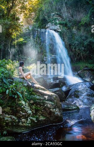 Eine schöne junge Frau, die in einem Sonnenstrahl vor einem unberührten Wasserfall im Regenwald, Brasilien, Südamerika sitzt Stockfoto