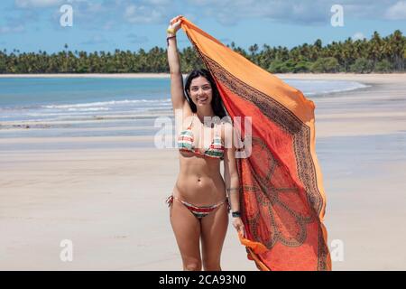 Eine schöne hispanische (lateinamerikanische) Frau in ihren Zwanzigern lächelnd, hält einen bunten Schal und steht an einem einsamen Strand, Brasilien, Südamerika Stockfoto