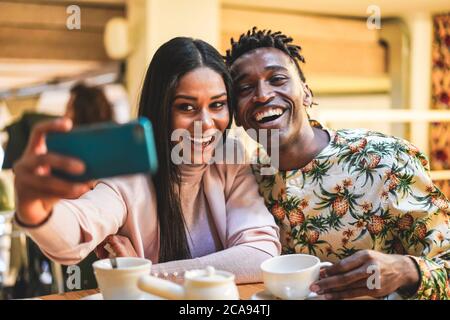 Glückliches schwarzes Paar, das Kaffee in der Bäckerei trinkt, während es Selfie mit dem Handy macht - Junge Leute, die Spaß mit Technologie-Trends haben - Social Media Stockfoto