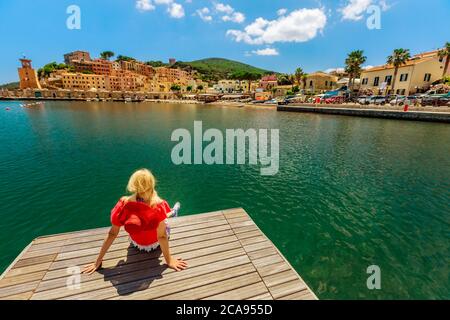 Blonde Frau in rotem Anzug Ruhe und Sonnenbaden auf Anlegesteg in Rio Marina Hafen der Insel Elba, Toskana, Italien, Europa Stockfoto