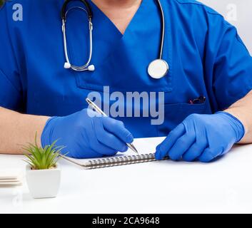 Der männliche Arzt in blauer Uniform sitzt an einem weißen Tisch und schreibt in einem Papiernotizbuch, mit sterilen Handschuhen an den Händen, dem Konzept, Patienten zu empfangen Stockfoto