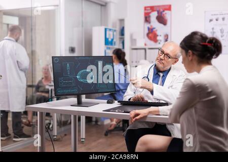 Leitender Arzt zeigt auf CT-Scan am Computer im Krankenhaus für junge Patienten. Junge Sanitäter diskutieren mit behinderten alten Frau im Rollstuhl auf Klinikkorridor. Krankenschwester trägt blaue Uniform. Stockfoto