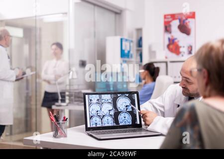 Der Radiologe zeigt auf den CT-Scan des Gehirns auf dem Laptop-Bildschirm während der Untersuchung einer älteren Frau im Krankenhausbüro. Reifer Arzt im weißen Mantel Diskussion mit kranken jungen Frau im Klinikkorridor. Stockfoto