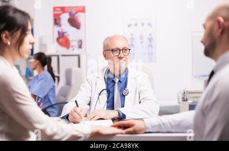 Besorgtes Paar, das während der medizinischen Untersuchung im Krankenhausbüro die Hände hielt. Leitender Arzt mit grauem Haar mit weißem Mantel und Stethoskop während der Untersuchung. Stockfoto