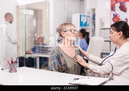 Arzt hören ältere Frau Herzschlag mit Stethoskop im Krankenhaus Büro trägt weißen Mantel. Behinderter älterer Mann im Rollstuhl, der mit einem Arzt auf dem Klinikkorridor diskutiert. Stockfoto