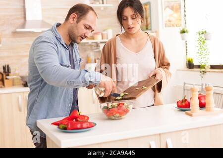 Mann gießt Olivenöl auf Salat in der Küche. Gesunder Salat mit frischem Gemüse. Glücklich in der Liebe fröhliche und sorglose Paar hilft einander, Mahlzeit vorzubereiten Stockfoto