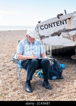 Aldeburgh, Suffolk, Großbritannien. August 2020. Ein Künstler ist zufrieden, am Kiesstrand an der Nordsee zu sitzen und zu zeichnen. Es war ein warmer Sommertag mit einer steifen Brise, da der aktuelle heiße Zauber im Osten Englands weitergeht. Kredit: Julian Eales/Alamy Live Nachrichten Stockfoto