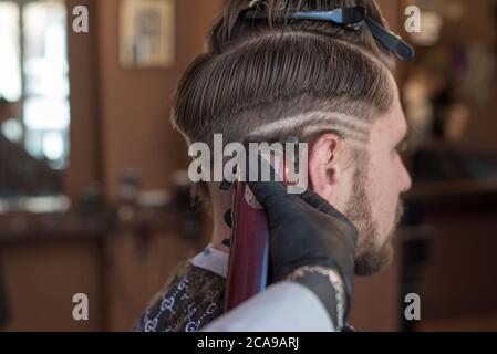 Ein Friseur schneidet einen bärtigen jungen Kerl mit einem Haarschneider und kämmt die Haare auf seinem Kopf. Die Arbeit des Meisters im Männerhaarschnitt in einem Friseurladen. Die Ba Stockfoto