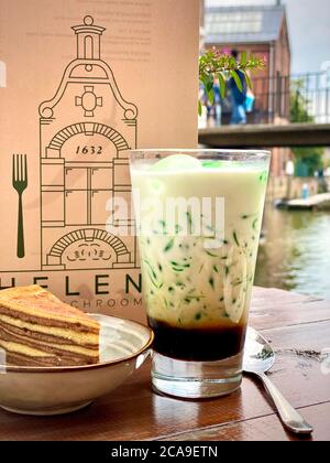 Traditionelles indonesisches süßes Erfrischungsgetränk Cendol aus Pandas-Saft, Mungbohne. Restaurant Cafe Holztisch draußen. Serviertes Getränk im Glas mit einem Stück Schichtkuchen in einer Schüssel mit einem Menü im Hintergrund. Bunt grün, braun cremig Getränk. Durstlöschender Durst. Stockfoto