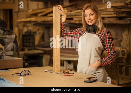 Selbstbewusste Holzarbeiterin Frau posiert vor der Kamera, halten Holzstück, tragen Uniform in der Werkstatt Stockfoto