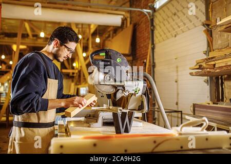 Selbstbewusste junge Tischler arbeiten mit Gehrungsäge in der Fabrik, stehen hinter einer Werkbank in Uniform. Industrie, Handwerk, Zimmerei Konzept Stockfoto