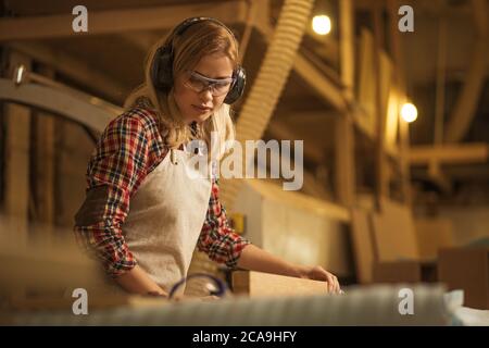 Konzentrierte seriöse, selbstbewusste Zimmerfrau beschäftigt sich mit Holzstücken, sie hört Musik, schützt ihre Ohren vor lauten Geräuschen in der Fabrik Stockfoto