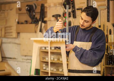 Der junge Möbelhersteller macht geschickt einen Holzstuhl auf einer Werkbank, während er allein in seiner großen Holzwerkstatt, Handwerk und Zimmerei concep arbeitet Stockfoto