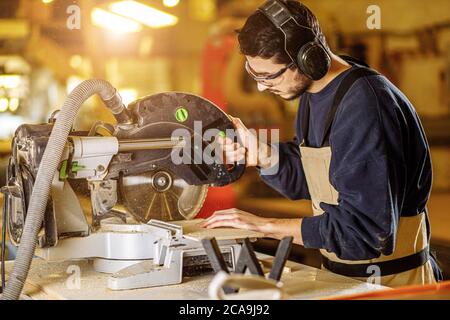 Seitenansicht auf konzentrierter kaukasischer Holzarbeiter, der ein Stück Holz auf einem kreisförmigen säger hinter der Werkbank sägt Stockfoto