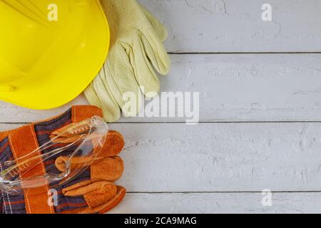 Bauarbeit Sicherheitsschutzausrüstung mit gelbem Helm und ein Paar Arbeitshandschuhe Schutzbrille auf einem weißen Hintergrund aus Holz Stockfoto