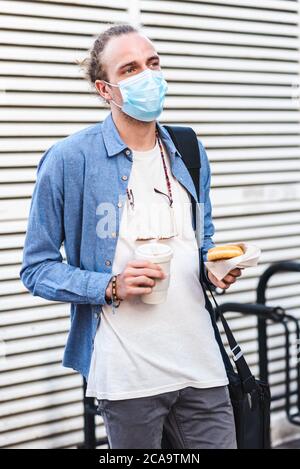 Junger Mann Dressing lässig mit chirurgischen Maske und hält in den Händen eine Kaffee Thermoskanne und Donut Stockfoto