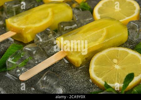 Nahaufnahme gefrorener Fruchtsaftpopsicles mit frischen Zitronenscheiben, grünen Minzblättern und Eiswürfeln auf grauer Tischfläche, Hochwinkelansicht Stockfoto