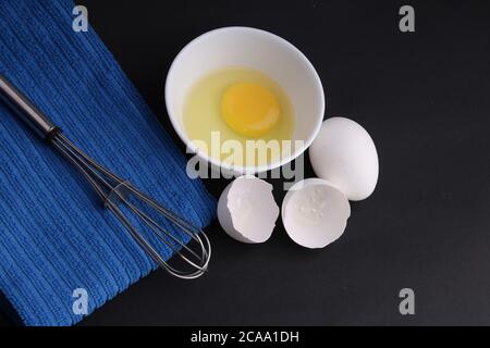 Zerbrochenes Hühnerei mit schwarzem Pfeffer. Es ist bereit, mit Whisker behackt zu werden. Es ist auf schwarzem Hintergrund mit blau gestreiften Servietten gehalten. Stockfoto