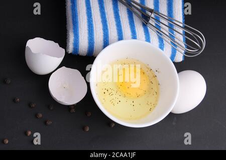 Zerbrochenes Hühnerei mit schwarzem Pfeffer. Es ist bereit, mit Whisker behackt zu werden. Es ist auf schwarzem Hintergrund mit blau gestreiften Servietten gehalten. Stockfoto