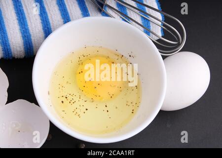 Zerbrochenes Hühnerei mit schwarzem Pfeffer. Es ist bereit, mit Whisker behackt zu werden. Es ist auf schwarzem Hintergrund mit blau gestreiften Servietten gehalten. Stockfoto